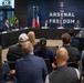 A formal roundtable meeting is taking place at a venue branded 'The Arsenal of Freedom,' with several officials and attendees seated at a curved conference table. The setting includes American and Texas state flags, along with multiple military branch flags displayed in the background. An audience of approximately 20-25 people is seated facing the panel.
