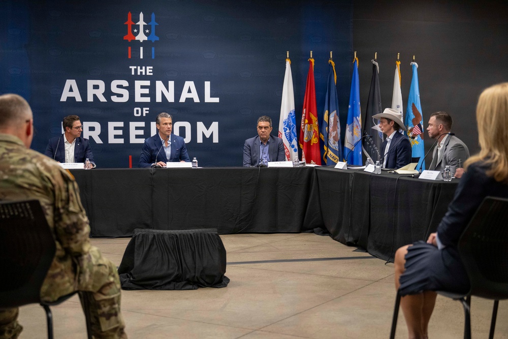 A formal panel discussion or briefing is taking place at a venue branded 'The Arsenal of Freedom,' with five individuals seated at a long table covered in black cloth. Multiple U.S. military branch flags are displayed in the background. The audience includes at least one person in military camouflage uniform and a civilian woman.