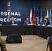 A formal panel discussion or briefing is taking place at a venue branded 'The Arsenal of Freedom,' with five individuals seated at a long table covered in black cloth. Multiple U.S. military branch flags are displayed in the background. The audience includes at least one person in military camouflage uniform and a civilian woman.