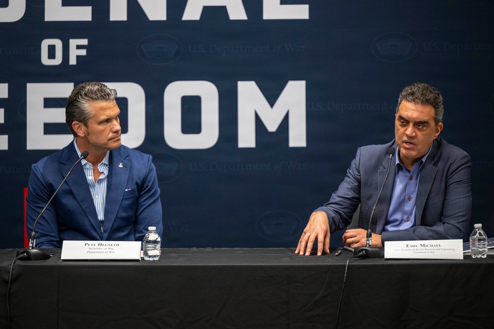 Two men are seated at a panel table with name placards identifying them as Pete Hegseth (Secretary of War, Department of War) and Emil Michael (Under Secretary of War for Research and Engineering, Department of War). They appear to be participating in a formal press conference or panel discussion in front of a large backdrop reading 'Arsenal of Freedom' with U.S. Department of War branding. The man on the right appears to be speaking while the man on the left listens.