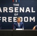 Three men in business attire are seated at a panel table in front of a large navy blue banner reading 'The Arsenal of Freedom' with 'U.S. Department of War' watermarks visible in the background. Name placards are visible in front of each participant, identifying them as Bryan Chambers, Pete Hegseth, and Eddie Michaels. The setting appears to be a formal press conference or panel discussion.