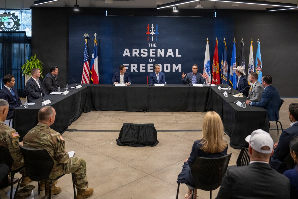 A formal roundtable meeting is taking place at a venue branded 'The Arsenal of Freedom,' with participants seated at a U-shaped table arrangement. Multiple individuals in business attire and at least one military uniform are present, with an audience seated facing the main table. The backdrop features the American flag, Texas state flag, and multiple U.S. military branch flags.