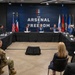 A formal roundtable meeting is taking place at a venue branded 'The Arsenal of Freedom,' with participants seated at a U-shaped table arrangement. Multiple individuals in business attire and at least one military uniform are present, with an audience seated facing the main table. The backdrop features the American flag, Texas state flag, and multiple U.S. military branch flags.