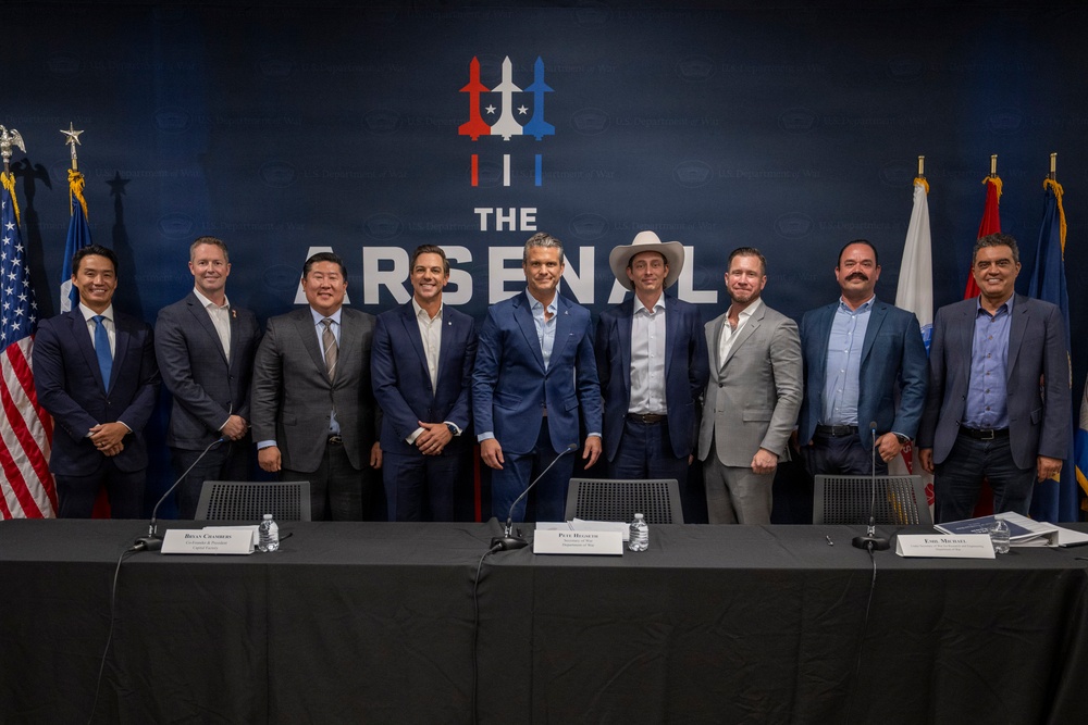 A group of nine men pose for a formal photo in front of a branded backdrop reading 'The Arsenal,' featuring a logo with aircraft and stars in red, white, and blue. The setting appears to be an official press conference or signing event, with a draped table in the foreground containing name placards, microphones, and water bottles. Flags including the American flag are visible on either side of the backdrop.