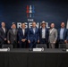 A group of nine men pose for a formal photo in front of a branded backdrop reading 'The Arsenal,' featuring a logo with aircraft and stars in red, white, and blue. The setting appears to be an official press conference or signing event, with a draped table in the foreground containing name placards, microphones, and water bottles. Flags including the American flag are visible on either side of the backdrop.