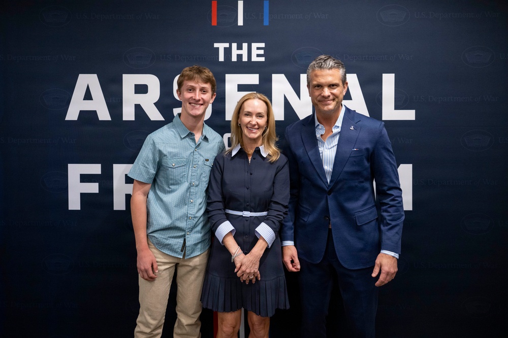 Three individuals are posing together in front of a dark navy backdrop that reads 'The Arsenal of Freedom' with a 'U.S. Department of War' watermark pattern. The group consists of a young man on the left, a woman in the center, and an older man on the right, all smiling for a formal photo opportunity. The setting appears to be an official or promotional event.