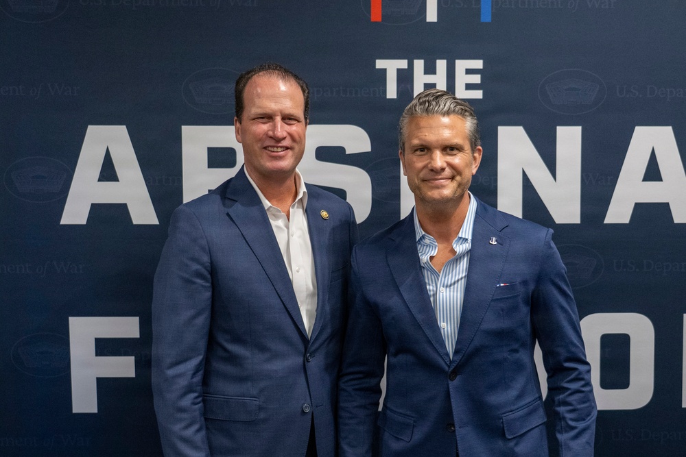 Two men in navy blue blazers are posing together in front of a branded backdrop reading 'The Arsenal F[orum/und]' with 'U.S. Department of War' logos visible in the background. Both men are smiling at the camera in what appears to be a formal or semi-formal event setting. The backdrop features red, white, and blue design elements consistent with a U.S. government or defense-related event.