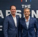 Two men in navy blue blazers are posing together in front of a branded backdrop reading 'The Arsenal F[orum/und]' with 'U.S. Department of War' logos visible in the background. Both men are smiling at the camera in what appears to be a formal or semi-formal event setting. The backdrop features red, white, and blue design elements consistent with a U.S. government or defense-related event.