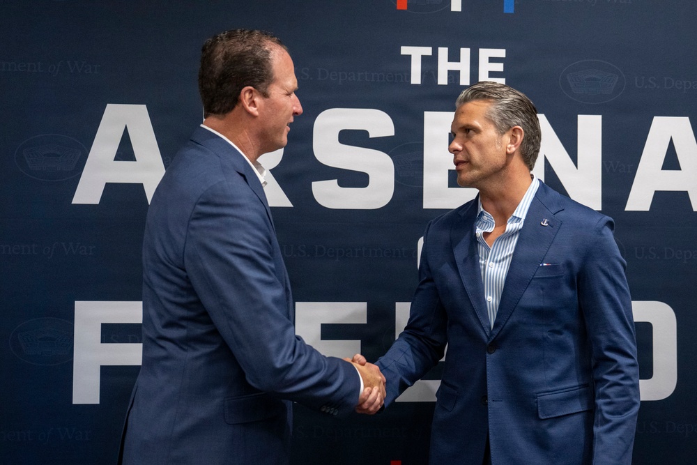 Two men in navy blue suits are shaking hands in front of a backdrop that reads 'The Arsenal of Freedom' with U.S. Department of Defense branding visible. The setting appears to be a formal event or press occasion. Both men are dressed professionally and appear engaged in a formal greeting or agreement.