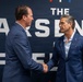 Two men in navy blue suits are shaking hands in front of a backdrop that reads 'The Arsenal of Freedom' with U.S. Department of Defense branding visible. The setting appears to be a formal event or press occasion. Both men are dressed professionally and appear engaged in a formal greeting or agreement.