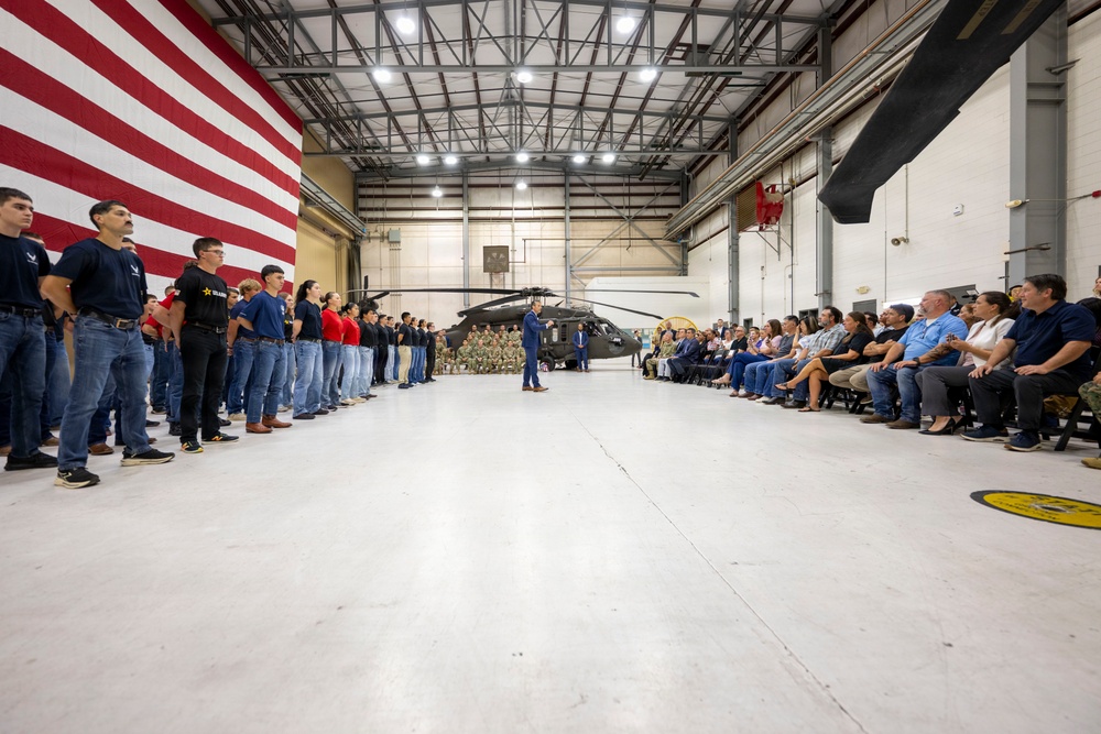 A formal ceremony is taking place inside a military aircraft hangar, with a large American flag displayed on the left wall and a military helicopter (appears to be a UH-60 Black Hawk) positioned in the background. A speaker in a blue suit stands at the center addressing two groups: uniformed and civilian youth standing in formation on the left, and seated adult attendees on the right. The event appears to be an official military or JROTC-related ceremony or recognition event.