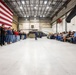 A formal ceremony is taking place inside a military aircraft hangar, with a large American flag displayed on the left wall and a military helicopter (appears to be a UH-60 Black Hawk) positioned in the background. A speaker in a blue suit stands at the center addressing two groups: uniformed and civilian youth standing in formation on the left, and seated adult attendees on the right. The event appears to be an official military or JROTC-related ceremony or recognition event.