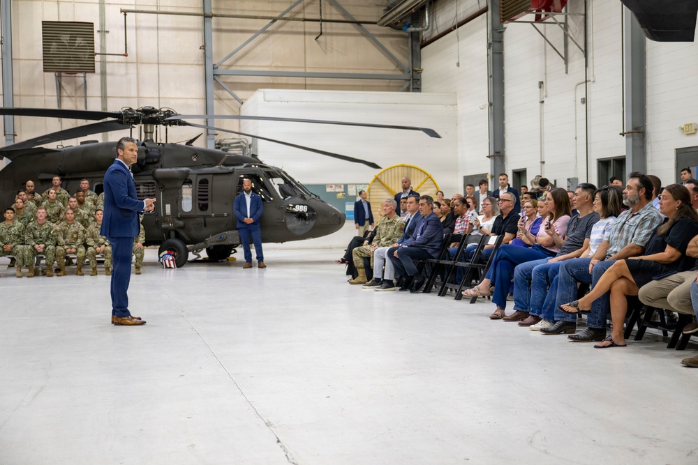 A civilian in a blue suit is addressing a mixed audience of military personnel in camouflage uniforms and civilians seated in folding chairs inside a military hangar. A dark military helicopter, appearing to be a UH-60 Black Hawk with tail number 986, is prominently displayed in the background. The setting suggests an official briefing, town hall, or ceremonial event at a military installation.