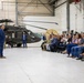 A civilian in a blue suit is addressing a mixed audience of military personnel in camouflage uniforms and civilians seated in folding chairs inside a military hangar. A dark military helicopter, appearing to be a UH-60 Black Hawk with tail number 986, is prominently displayed in the background. The setting suggests an official briefing, town hall, or ceremonial event at a military installation.