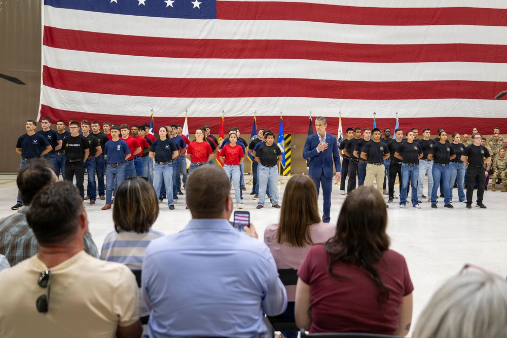 A military enlistment or swearing-in ceremony is taking place inside what appears to be a large hangar or military facility. A group of young recruits wearing various branch-branded t-shirts (Army, Air Force, Marines) and jeans stand in formation before a large American flag backdrop, while a man in a blue suit stands among them with his hand over his heart. An audience of family members and observers watches from seated positions in the foreground.