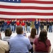 A military enlistment or swearing-in ceremony is taking place inside what appears to be a large hangar or military facility. A group of young recruits wearing various branch-branded t-shirts (Army, Air Force, Marines) and jeans stand in formation before a large American flag backdrop, while a man in a blue suit stands among them with his hand over his heart. An audience of family members and observers watches from seated positions in the foreground.