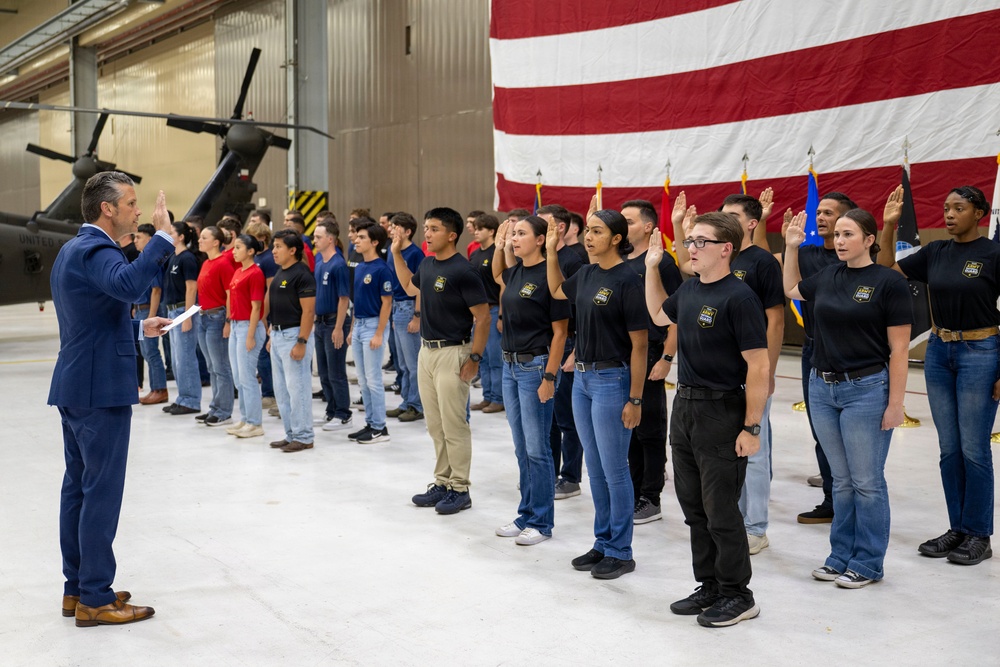 A formal military enlistment or oath-of-service ceremony is taking place inside a military hangar. A man in a blue suit stands facing a large group of young recruits who have their right hands raised, taking an oath. A large American flag serves as a backdrop, with military helicopters and unit colors visible in the background.