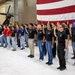 A formal military enlistment or oath-of-service ceremony is taking place inside a military hangar. A man in a blue suit stands facing a large group of young recruits who have their right hands raised, taking an oath. A large American flag serves as a backdrop, with military helicopters and unit colors visible in the background.