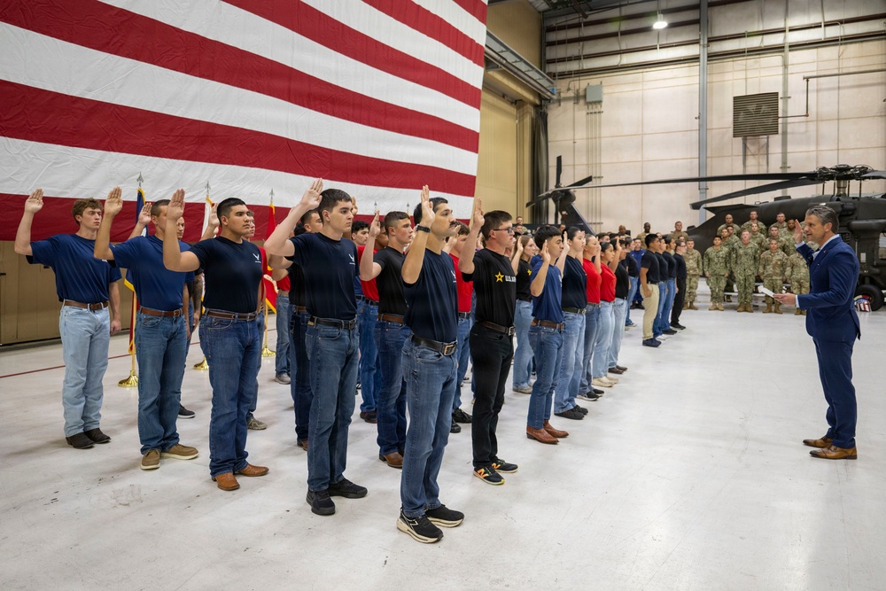 A military enlistment or oath-of-enlistment ceremony is taking place inside a large aircraft hangar. A group of approximately 30 civilians in casual clothing (jeans and colored t-shirts) are raising their right hands while a man in a blue suit administers the oath. A large American flag serves as a backdrop, with uniformed military personnel observing from the right side.