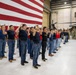 A military enlistment or oath-of-enlistment ceremony is taking place inside a large aircraft hangar. A group of approximately 30 civilians in casual clothing (jeans and colored t-shirts) are raising their right hands while a man in a blue suit administers the oath. A large American flag serves as a backdrop, with uniformed military personnel observing from the right side.