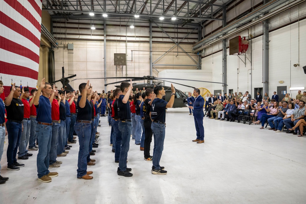 A military enlistment or oath-of-service ceremony is taking place inside a large aircraft hangar. A group of young recruits in civilian clothing are raising their right hands while a man in a blue suit administers the oath. A large American flag, a military helicopter, and an audience of observers are visible in the background.