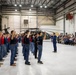 A military enlistment or oath-of-service ceremony is taking place inside a large aircraft hangar. A group of young recruits in civilian clothing are raising their right hands while a man in a blue suit administers the oath. A large American flag, a military helicopter, and an audience of observers are visible in the background.