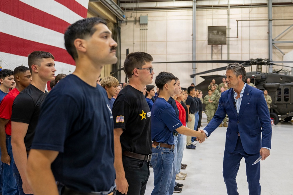 A man in a blue suit is shaking hands with young individuals lined up in a military hangar facility. The group appears to consist of youth recruits or cadets wearing casual civilian clothing with some Army-branded shirts. Military personnel in camouflage uniforms and a Black Hawk helicopter are visible in the background.