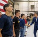 A man in a blue suit is shaking hands with young individuals lined up in a military hangar facility. The group appears to consist of youth recruits or cadets wearing casual civilian clothing with some Army-branded shirts. Military personnel in camouflage uniforms and a Black Hawk helicopter are visible in the background.