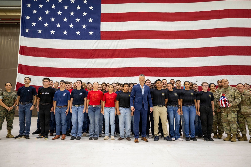A large group of young people, likely military recruits or JROTC cadets, pose for a group photo in front of an oversized American flag inside what appears to be a military hangar or large indoor facility. A man in a blue suit stands centrally among the group, flanked by individuals in military camouflage uniforms on the far sides. The group wears a mix of branded military branch t-shirts (U.S. Army, Air Force, Marines) and civilian clothing.