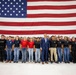 A large group of young people, likely military recruits or JROTC cadets, pose for a group photo in front of an oversized American flag inside what appears to be a military hangar or large indoor facility. A man in a blue suit stands centrally among the group, flanked by individuals in military camouflage uniforms on the far sides. The group wears a mix of branded military branch t-shirts (U.S. Army, Air Force, Marines) and civilian clothing.