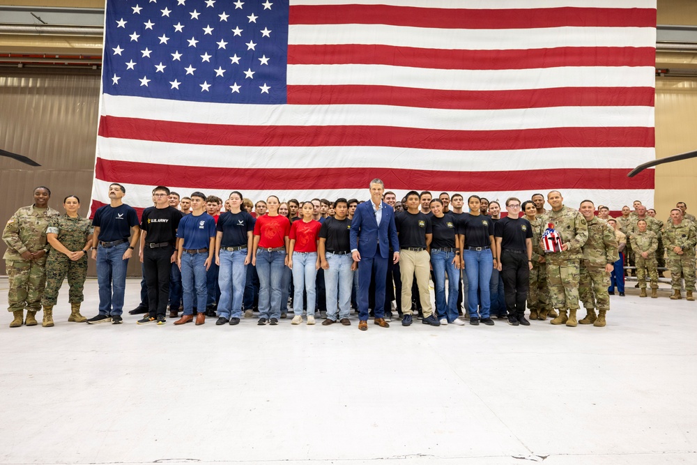 A large group photo taken inside what appears to be a military hangar, featuring uniformed military personnel flanking a group of younger individuals in civilian/JROTC-style attire. A man in a blue suit stands at the center of the group. A large American flag serves as the backdrop, spanning nearly the entire width of the frame.