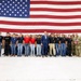 A large group photo taken inside what appears to be a military hangar, featuring uniformed military personnel flanking a group of younger individuals in civilian/JROTC-style attire. A man in a blue suit stands at the center of the group. A large American flag serves as the backdrop, spanning nearly the entire width of the frame.