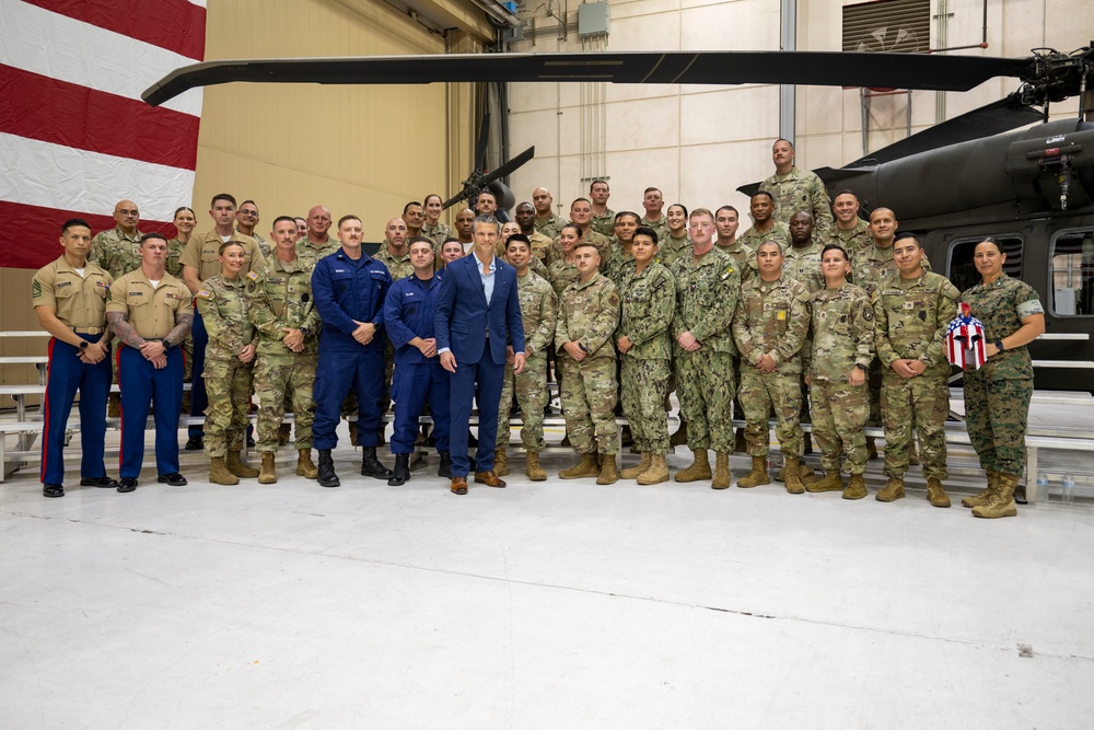 A large group of approximately 40-50 military personnel from multiple U.S. service branches pose for a formal group photograph inside a military aircraft hangar. One civilian in a blue suit stands prominently in the center of the group. Black military helicopters and a large American flag are visible in the background.