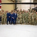 A large group of approximately 40-50 military personnel from multiple U.S. service branches pose for a formal group photograph inside a military aircraft hangar. One civilian in a blue suit stands prominently in the center of the group. Black military helicopters and a large American flag are visible in the background.