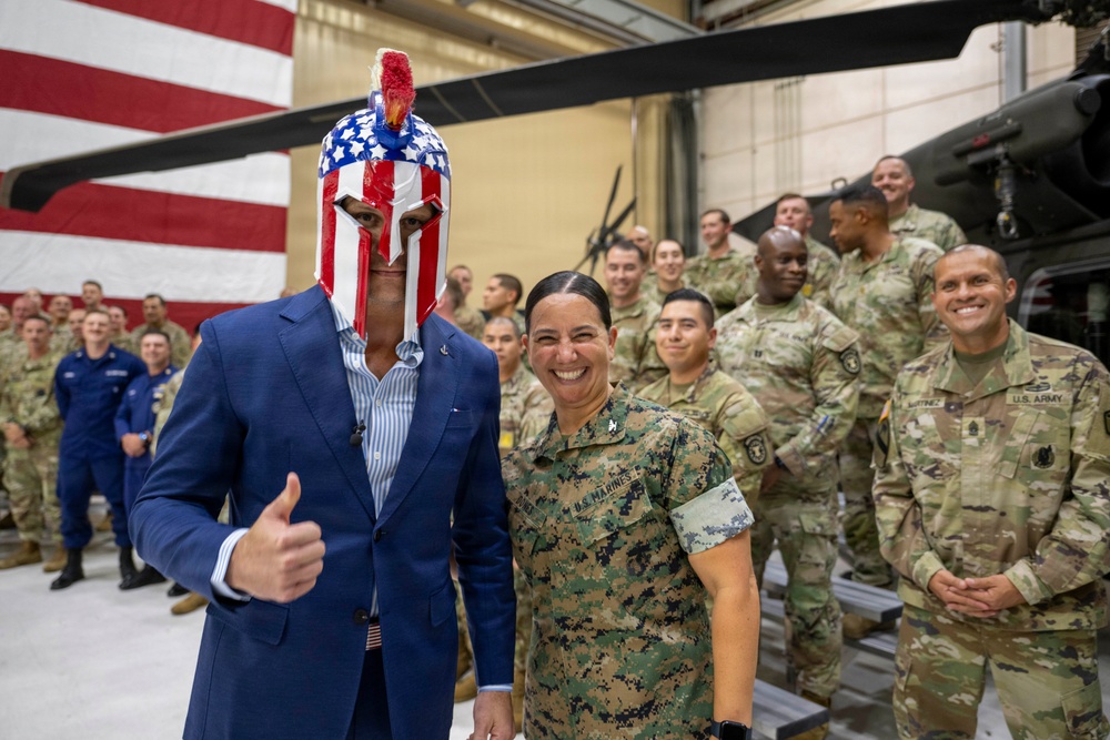 A civilian in a blue suit wearing a patriotic American flag-themed Spartan helmet gives a thumbs-up while posing with military personnel in a hangar setting. A U.S. Marines service member stands beside him smiling broadly, with a large group of Army and other branch personnel visible in the background. A large American flag and military helicopters are visible in the hangar.