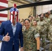 A civilian in a blue suit wearing a patriotic American flag-themed Spartan helmet gives a thumbs-up while posing with military personnel in a hangar setting. A U.S. Marines service member stands beside him smiling broadly, with a large group of Army and other branch personnel visible in the background. A large American flag and military helicopters are visible in the hangar.