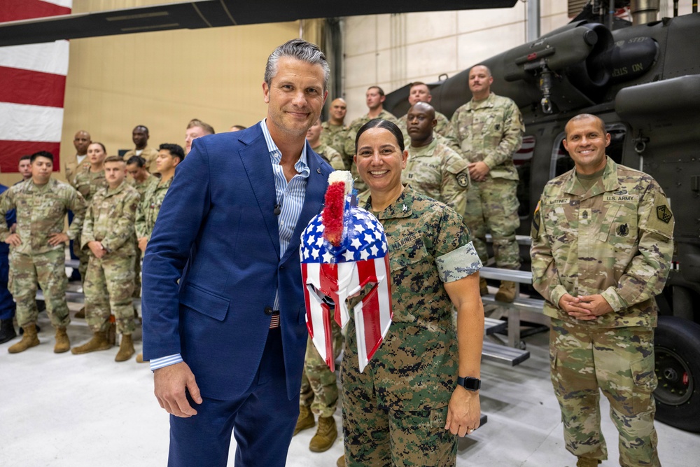 A civilian man in a blue suit poses alongside a female Marine officer in a military hangar, with a group of U.S. Army soldiers standing in formation behind them. The Marine officer is holding a patriotic American flag-themed decorative item. A military helicopter is partially visible in the background.