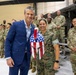 A civilian man in a blue suit poses alongside a female Marine officer in a military hangar, with a group of U.S. Army soldiers standing in formation behind them. The Marine officer is holding a patriotic American flag-themed decorative item. A military helicopter is partially visible in the background.