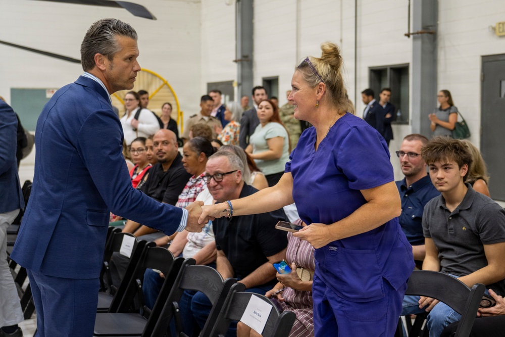 A man in a blue suit is shaking hands with a woman wearing blue medical scrubs at what appears to be a formal gathering or ceremony held in a large indoor facility. The woman is holding what appears to be a small award or recognition item. A crowd of seated and standing attendees is visible in the background.