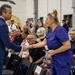 A man in a blue suit is shaking hands with a woman wearing blue medical scrubs at what appears to be a formal gathering or ceremony held in a large indoor facility. The woman is holding what appears to be a small award or recognition item. A crowd of seated and standing attendees is visible in the background.