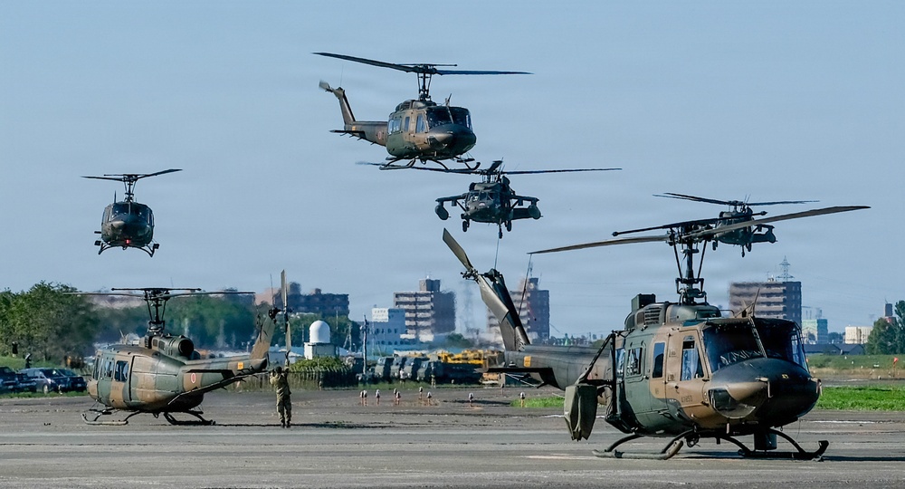 A military airfield scene showing multiple helicopters in various stages of operation, including aircraft on the ground and several airborne. The helicopters appear to be UH-1 Iroquois (Huey) variants and what appears to be a UH-60 Black Hawk, painted in olive drab military camouflage. A ground crew member is visible on the tarmac, and an urban skyline is visible in the background.