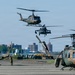 A military airfield scene showing multiple helicopters in various stages of operation, including aircraft on the ground and several airborne. The helicopters appear to be UH-1 Iroquois (Huey) variants and what appears to be a UH-60 Black Hawk, painted in olive drab military camouflage. A ground crew member is visible on the tarmac, and an urban skyline is visible in the background.