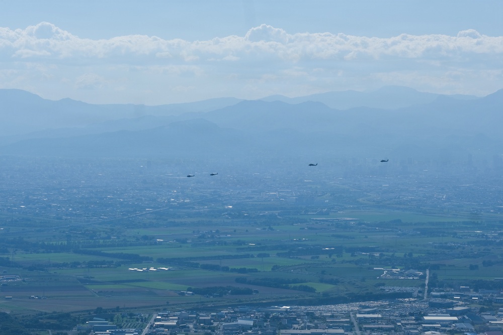 An aerial or elevated view of a wide urban and agricultural valley with mountain ranges visible in the hazy background. Four dark aircraft, appearing to be helicopters, are flying in a loose formation across the middle distance. The scene is characterized by atmospheric haze reducing visibility across the landscape.