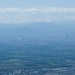 An aerial or elevated view of a wide urban and agricultural valley with mountain ranges visible in the hazy background. Four dark aircraft, appearing to be helicopters, are flying in a loose formation across the middle distance. The scene is characterized by atmospheric haze reducing visibility across the landscape.