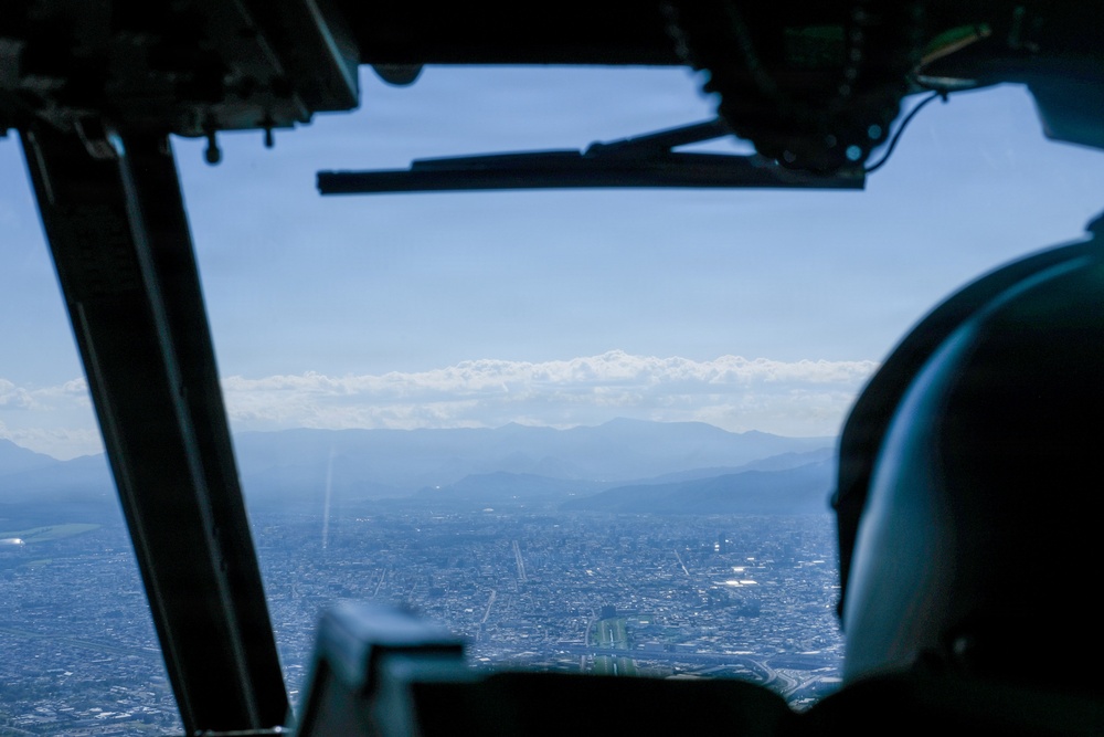 The image is taken from inside a military or transport aircraft cockpit, looking out through the windshield at a sprawling urban area below. In the background, a mountain range with snow-capped peaks is visible under a hazy blue sky. A helmeted crew member is partially visible in the right foreground.