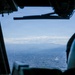 The image is taken from inside a military or transport aircraft cockpit, looking out through the windshield at a sprawling urban area below. In the background, a mountain range with snow-capped peaks is visible under a hazy blue sky. A helmeted crew member is partially visible in the right foreground.