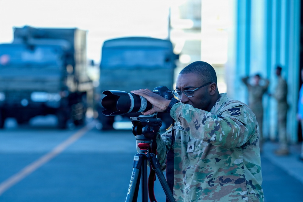 A military photographer in OCP (Operational Camouflage Pattern) uniform is actively shooting with a Canon DSLR camera mounted on a Manfrotto tripod. The setting appears to be a military airfield or base, with military trucks visible in the blurred background. Additional personnel in uniform can be seen standing near a structure in the background.