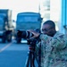 A military photographer in OCP (Operational Camouflage Pattern) uniform is actively shooting with a Canon DSLR camera mounted on a Manfrotto tripod. The setting appears to be a military airfield or base, with military trucks visible in the blurred background. Additional personnel in uniform can be seen standing near a structure in the background.