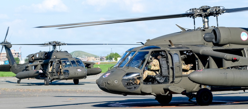 The image shows multiple military helicopters, identifiable as UH-60 Black Hawks, positioned on a tarmac or airfield. The helicopters are painted in olive drab/dark green military livery and are crewed by personnel in military flight gear. The setting appears to be a military airfield with urban and hilly terrain visible in the background.