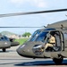 The image shows multiple military helicopters, identifiable as UH-60 Black Hawks, positioned on a tarmac or airfield. The helicopters are painted in olive drab/dark green military livery and are crewed by personnel in military flight gear. The setting appears to be a military airfield with urban and hilly terrain visible in the background.